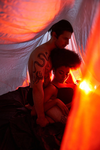 sexy multicultureel paar zitten onder lakens en poseren liefdevol omgeven door lichten, samen - Foto, afbeelding