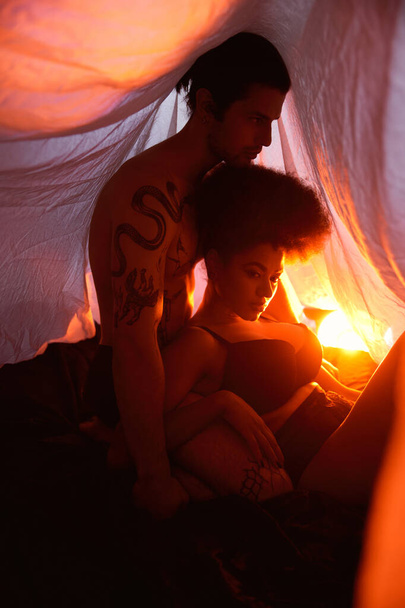 mladý rozmanitý pár sedící pod prostěradlem a pózující láskyplně obklopený světly, - Fotografie, Obrázek