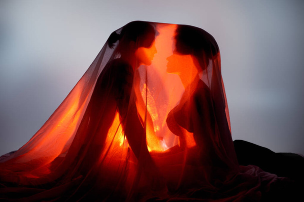 multicultural sexy couple covered with bedsheet surrounded by light on gray background, togetherness - Foto, Imagen