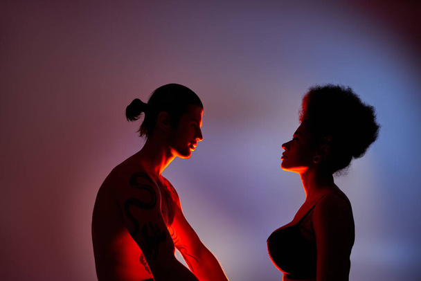 lákavé multikulturní pár dívá na sebe láskyplně na šedém pozadí se světly - Fotografie, Obrázek