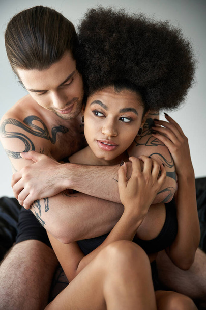 молодая привлекательная мультикультурная пара в нижнем белье сидит на кровати и обнимается с любовью - Фото, изображение