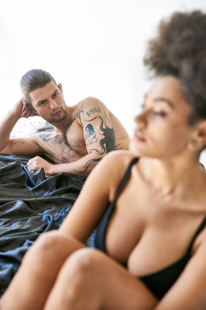 concentrarsi su bello uomo con tatuaggi sdraiato a letto accanto alla sua ragazza afroamericana offuscata - Foto, immagini