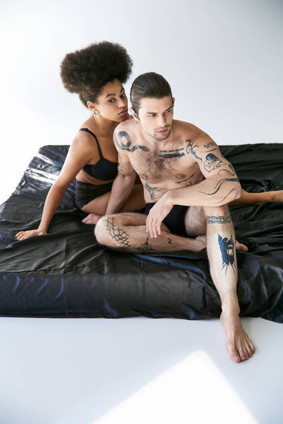 goed uitziende man met tatoeages zitten op bed naast zijn afrikaanse amerikaanse vriendin, sexy paar - Foto, afbeelding