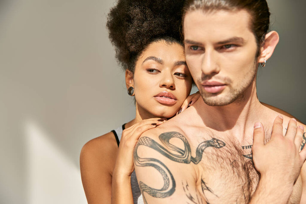szexi fiatal férfi tetoválásokkal átöleli gyönyörű afro-amerikai barátnője szürke háttér - Fotó, kép
