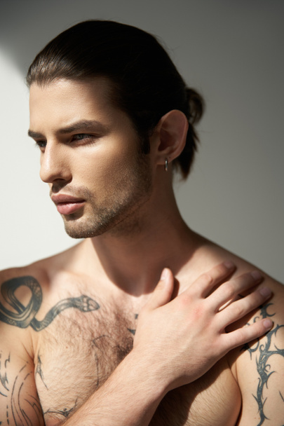 привлекательный молодой человек с татуировками и хвостиком, позирующий рукой на груди и отворачивающийся - Фото, изображение