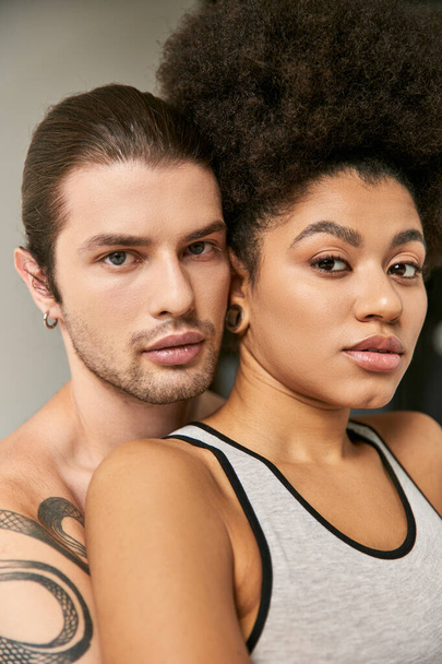 young tattooed man hugging his beautiful african american girlfriend on gray backdrop, sexy couple - Photo, Image