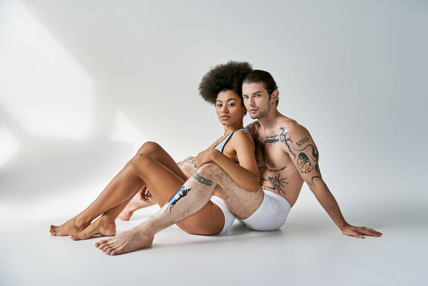 gut aussehendes sexy multikulturelles Paar in bequemer Unterwäsche auf dem Boden sitzend und liebevoll umarmt - Foto, Bild