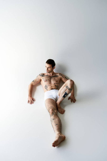 Вид сверху на красивого молодого человека с хвостиком и татуировками в нижнем белье, лежащего на полу очаровательно - Фото, изображение