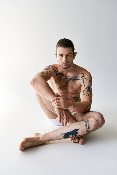 goed uitziende jonge getatoeëerde man model in ondergoed zitten op de vloer en kijken naar camera - Foto, afbeelding