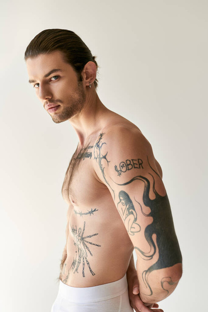δελεαστικός άνθρωπος με αλογοουρά και κομψά τατουάζ σε άνετο εσώρουχα ποζάρουν σε εκρού φόντο - Φωτογραφία, εικόνα