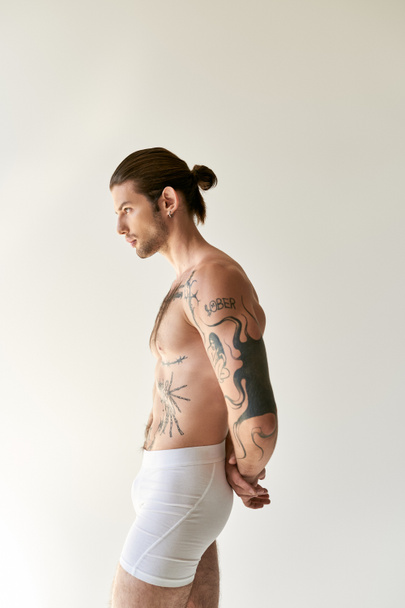 mladý dobře vypadající muž s culíkem a cool tetování ve spodním prádle pózování na ecru pozadí - Fotografie, Obrázek