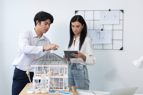 Gyönyörű fiatal kaukázusi projektmenedzser elmondja ázsiai mérnök épület részletesen segítségével tabletta, míg a mérnök mérési ház modell tárgyalóasztal tervrajz szórás körül. Szeplőtelen - Fotó, kép