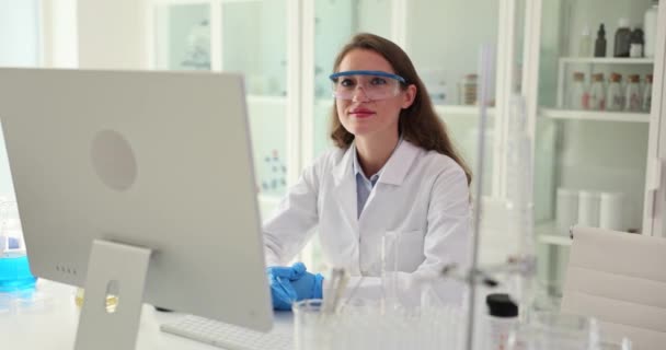 La química femenina concentrada en gafas de seguridad se sienta a la mesa con la computadora y los especímenes para experimentos. Trabajos de laboratorio e investigación en cámara lenta - Metraje, vídeo