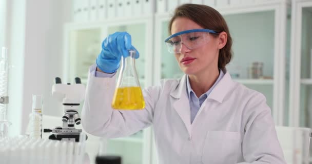 Trabalhadora do laboratório em óculos de segurança detém amostra de reagente na clínica leve. Cientista estuda propriedades químicas líquidas câmera lenta - Filmagem, Vídeo