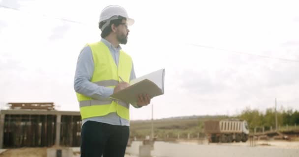 Man engineer in protective helmet and vest uniform standing at construction site, checking process of building and writing notes in notepad. Male inspector working at architect project outdoors - Footage, Video
