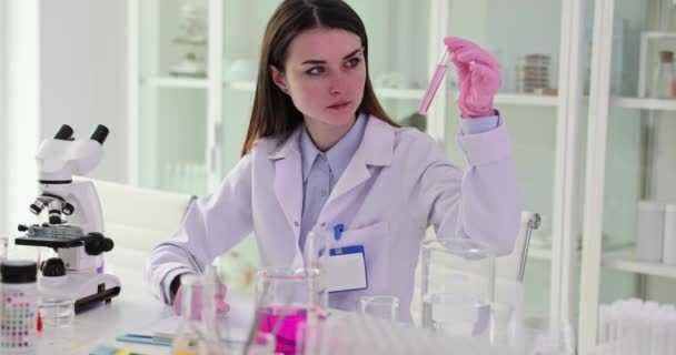Científico describe el color y el estado del reactivo químico examinado en el laboratorio. Mujer en guantes de goma sostiene tubo con líquido rosa sentado en la mesa de cámara lenta - Metraje, vídeo