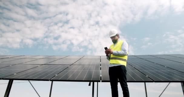 Low angle view on man engineer in uniform and protective helmet using cellphone for monitoring photovoltaic panel system producing clean renewable solar energy against sky background - Footage, Video