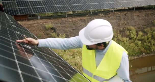屋外の太陽光発電所で太陽電池パネルを点検するベストおよび保護ヘルメットのエンジニアの人. ソーラーファームで働く男性スペシャリスト. 代替エネルギー源とイノベーションの概念 - 映像、動画