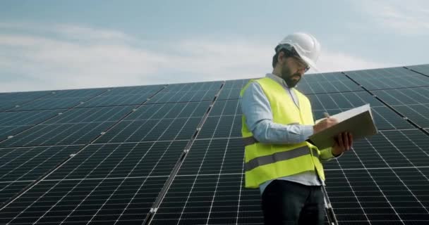 Egyenruhás munkás és védősisak áll a fotovoltaikus erőműben, vizsgálja a napelemeket a szabadban, és jegyzetel. A napelemes gazdaság karbantartása. - Felvétel, videó