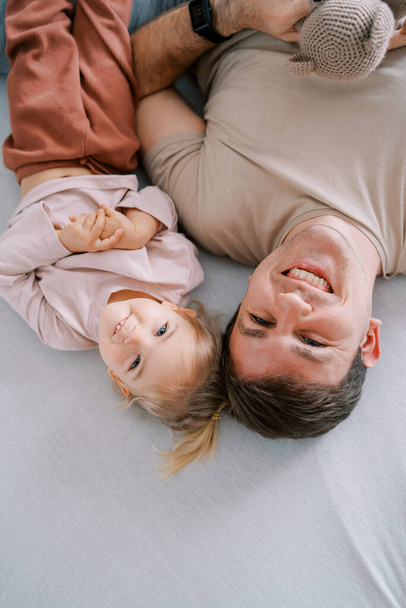O pai sorridente com uma menina deita-se de cabeça para baixo na cama. Vista superior. Foto de alta qualidade - Foto, Imagem