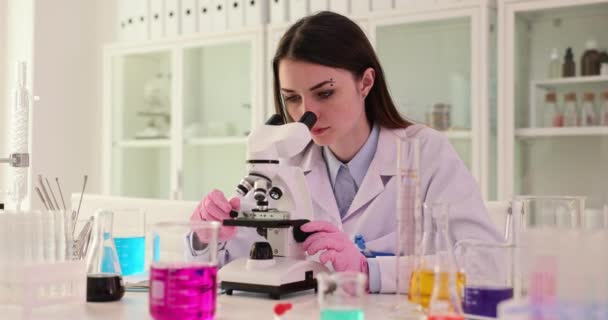 Aandachtige vrouwelijke wetenschapper kijkt naar monster in microscoop zittend op de werkplek. Medische faciliteit voor het testen met apparatuur voor het uitvoeren van experimenten slow motion - Video