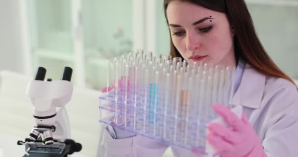 A koncentrált női vegyész megvizsgálja a csövek sorait az eszközökkel felszerelt laboratóriumban lévő mintákkal. A védőkesztyűs munkás kutatásokat végez a klinikán. - Felvétel, videó
