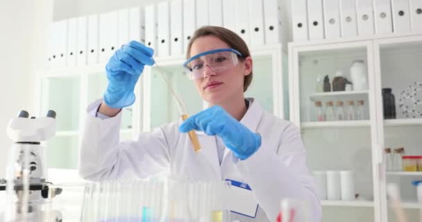 Femmina chimica in uniforme da lavoro mescola liquido in tubo di vetro con bastone esaminando campione a tavola con microscopio. Lavoro professionale in laboratorio - Filmati, video