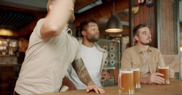 Młodzi mężczyźni piją piwo i oglądają mecz sportowy w pubie. Wesoły samiec świętuje bramkę i śmieje się z przyjaciół, ponieważ ich drużyna przegrywa mecz piłki nożnej - Materiał filmowy, wideo