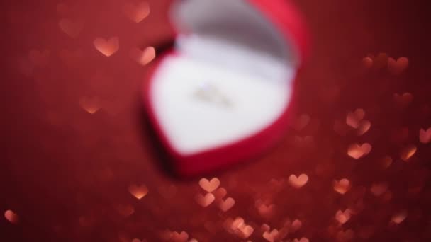 El anillo de compromiso se encuentra en una elegante caja sobre un fondo rojo con un recubrimiento de purpurina. Este fondo brilla con rayos de luz, creando un brillo encantador que da el anillo de compromiso incluso - Metraje, vídeo