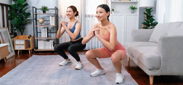Vigorous energetic woman with trainer or workout buddy doing exercise at home. Young athletic asian woman strength and endurance training session as home workout routine with squat. - Foto, Imagen