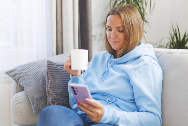 Una donna si rilassa su un comodo divano, smartphone in mano e una tazza di tè, abbracciando una perfetta miscela di tecnologia e tranquillità. - Foto, immagini