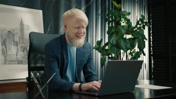 Mosolygó albínó vállalkozó jó híreket olvas a laptop képernyőjén. Boldog vezérigazgató elégedettnek érzi magát az új projekt a munkahelyen. Vidám üzletember, aki PC-t használ az irodában. Férfi szabadúszó élvezi a munkanapot  - Felvétel, videó