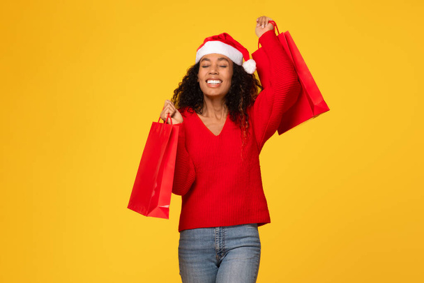 Χαρούμενη μαύρη γυναίκα φορώντας το καπέλο του Άη Βασίλη, κρατώντας κόκκινες τσάντες, προτείνοντας επιτυχημένες πωλήσεις Χριστουγέννων, στέκεται πάνω σε κίτρινο φόντο - Φωτογραφία, εικόνα