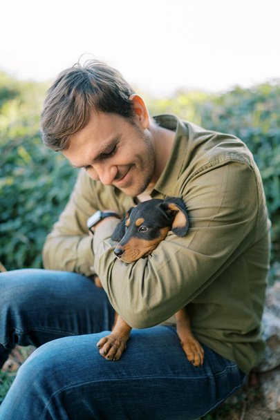 Jonge glimlachende man zit met een puppy op zijn knieën knuffelend en kijkend naar hem. Hoge kwaliteit foto - Foto, afbeelding