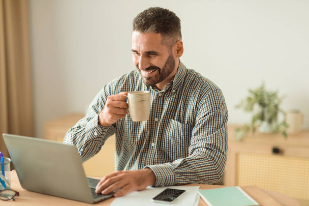 Feliz hombre de negocios de mediana edad que bebe café mientras usa el ordenador portátil en el lugar de trabajo en interiores, sentado durante el descanso con una taza de bebida caliente en el interior moderno de la oficina en casa. Emprendimiento exitoso - Foto, imagen