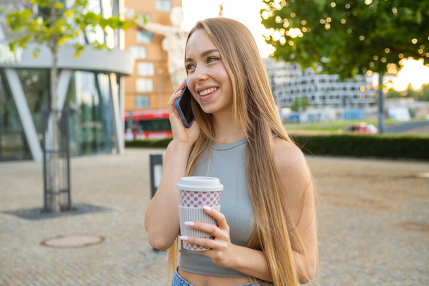 Happy young woman with blonde long loose hair spends time in park talking to boyfriend on phone. Lady cheerfully laughs drinking coffee outdoors - Photo, Image