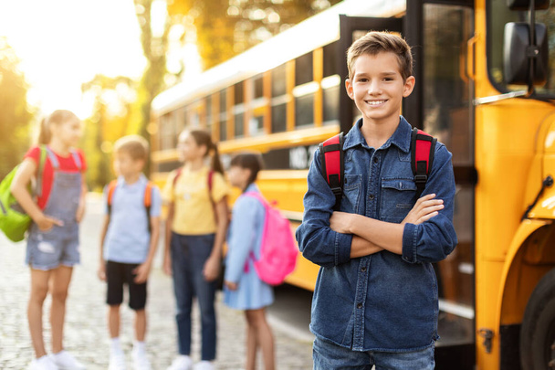 Χαμογελώντας Preteen αγόρι στέκεται με διπλωμένα τα χέρια κοντά στο κίτρινο σχολικό λεωφορείο και κοιτάζοντας κάμερα, ομάδα των συμμαθητών του κουβεντιάζοντας και γελώντας στο παρασκήνιο, τα παιδιά είναι έτοιμα για τη νέα ημέρα μάθησης - Φωτογραφία, εικόνα