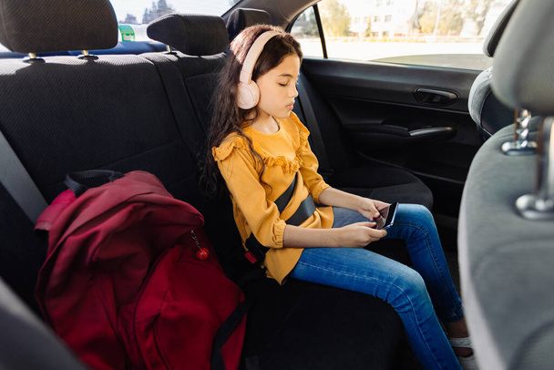 Hareket halindeki arabanın arka koltuğunda, odaklanmış genç bir kız kulaklık takıyor ve kendini akıllı telefona kaptırmış, yanında kırmızı okul çantası ve dışarıdaki dünya ile birlikte. - Fotoğraf, Görsel