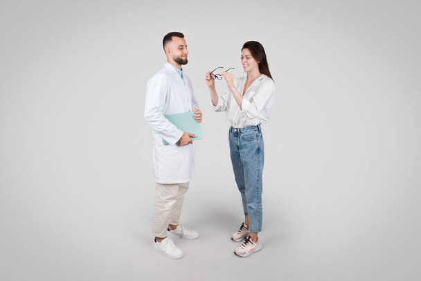 Giovane oftalmologo maschile europeo in camice bianco fornisce consulenza medica al paziente femminile, su sfondo grigio. colpo a tutta lunghezza - Foto, immagini