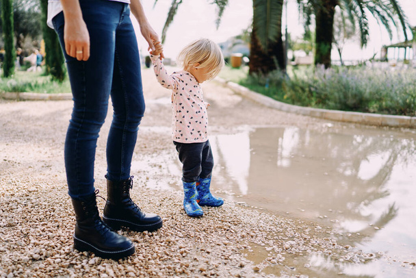 Mam houdt de hand vast van een klein meisje in rubberen laarzen, staande op de rand van een grote plas. Hoge kwaliteit foto - Foto, afbeelding
