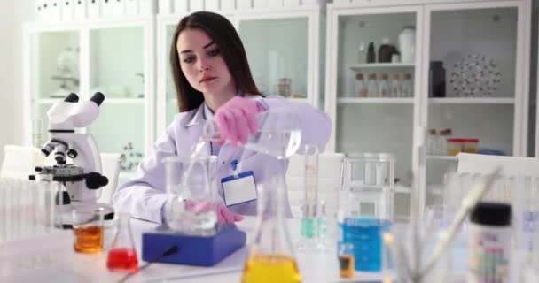 Женщина-ученый наливает прозрачную жидкость из стекла во фляжку, сидя на рабочем месте с микроскопом в лаборатории. Открытие экспертной замедленной съемки - Кадры, видео