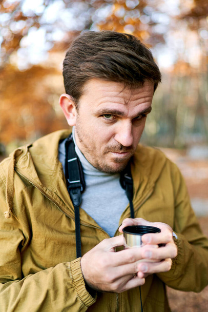 Νεαρός άνδρας με ένα φλιτζάνι καφέ στα χέρια του φαίνεται συνοφρυωμένος ενώ στέκεται στο πάρκο. Υψηλής ποιότητας φωτογραφία - Φωτογραφία, εικόνα