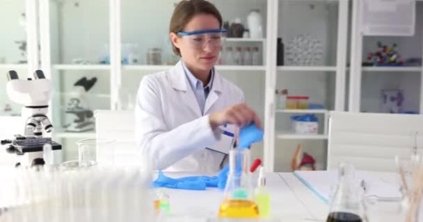 Donna scienziata indossa guanti di gomma prima di condurre l'esperimento seduto a tavola con microscopio e palloni. Professione importante e preparazione - Filmati, video