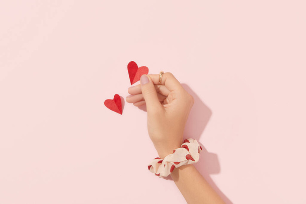 Ухоженная женская рука с розовыми матовыми ногтями, держащими бумажное сердце на розовом фоне. День святого Валентина концепция. Салон красоты, маникюр, педикюр. - Фото, изображение