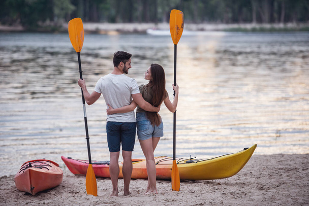Back view of young couple holding paddles and hugging while standing near the kayaks on the beach and watching sunset - Photo, Image