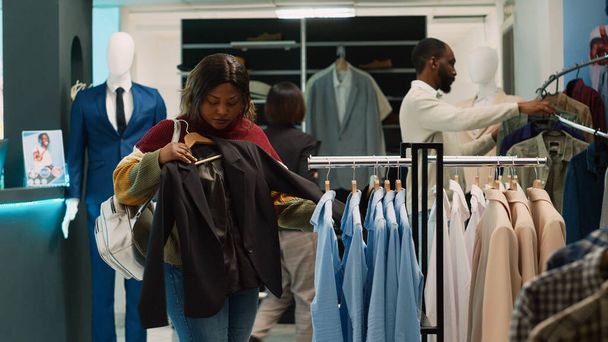 Mujer afroamericana tomando ropa formal de perchas para aumentar el guardarropa en la tienda de ropa. Joven cliente tratando de comprar nueva colección de ropa, analizando la tela en la boutique. Disparo de mano. - Foto, imagen