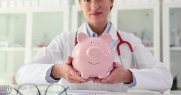 Vrouwelijke dokter verpleegster met roze spaarvarken in de kliniek. Ziektekostenverzekering gezondheidszorg en geneeskunde en ziekenhuisbudget en ziekenhuiskosten - Video