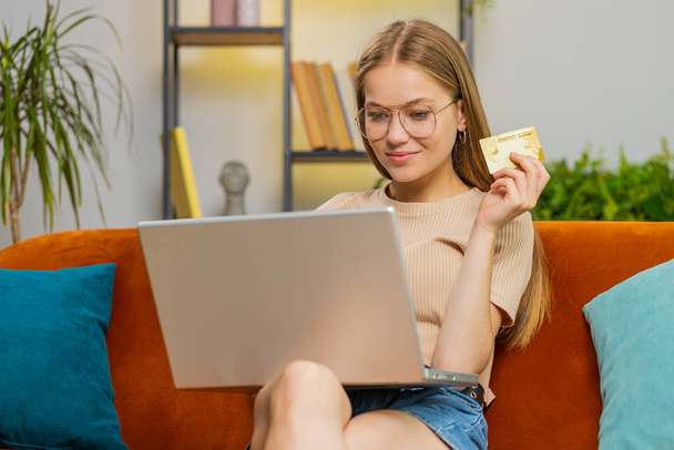 Young blonde woman using credit bank card and laptop computer notebook while transferring money, purchases online shopping, order food delivery at home. Adult girl in living room sitting on sofa couch - Photo, Image