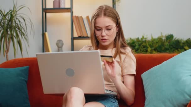 Junge blonde Frau mit Kreditkarte und Laptop, während sie Geld überweist, online einkauft, Essenslieferung zu Hause bestellt. Erwachsene Mädchen im Wohnzimmer sitzen auf Sofa-Couch - Filmmaterial, Video