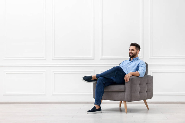 Handsome man sitting in armchair near white wall indoors, space for text - Foto, Bild